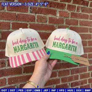 May include: Two baseball caps with off-white crowns and embroidered text reading bad day to be a MARGARITA. One cap features a pink and white striped brim, while the other has a solid green brim. Text at the top reads FLAT VERSION - 3 SIZE 4" 5" 6".