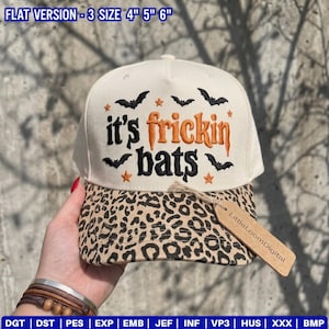 May include: A cream-colored baseball cap with a leopard print brim. The front features black and orange embroidered text that reads "it's frickin bats" surrounded by small black bat and orange star icons. A brown paper tag is attached to the brim.