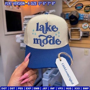 May include: A blue and beige trucker hat with the words "lake mode" embroidered on the front. The hat has a mesh back and a curved brim. A tag with the text "LittleLoomDigital" hangs from the hat.