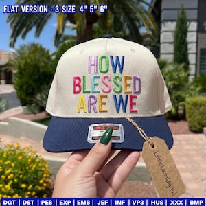 May include: A cream-colored baseball cap with a navy blue brim. The cap features the embroidered phrase "HOW BLESSED ARE WE" in colorful letters. A tag with the text "Little Loom Digital" hangs from the cap.
