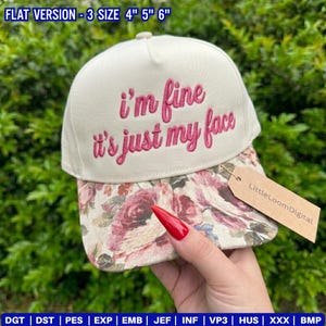 May include: A cream-colored baseball cap with the embroidered phrase "i'm fine it's just my face" in pink script. The brim features a floral pattern in shades of pink, red, and cream. A small tag with the text "LittleLoomDigital" is attached.