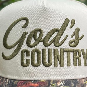 May include: A cream-coloured baseball cap with the embroidered text "God's COUNTRY" in olive green. The brim has a camouflage pattern. The cap is a classic, structured style.