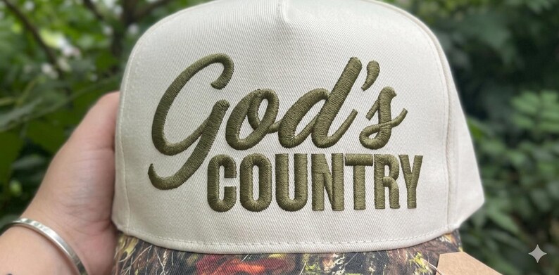 May include: A cream-coloured baseball cap with the embroidered text "God's COUNTRY" in olive green. The brim has a camouflage pattern. The cap is a classic, structured style.