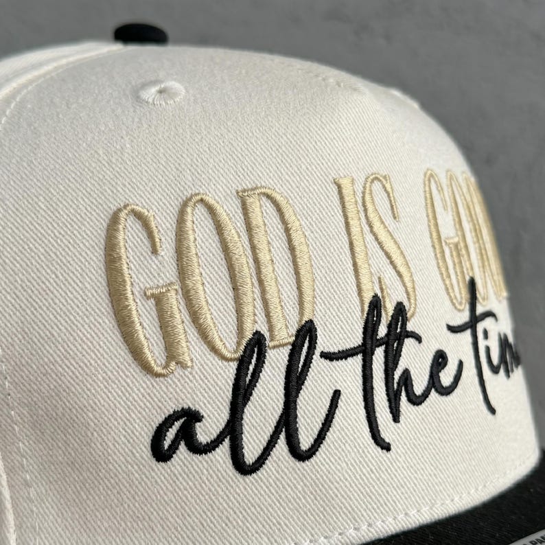 May include: A white and black baseball cap with the embroidered phrase "GOD IS GOOD all the time" in gold and black thread. The cap has a flat brim and a structured crown.