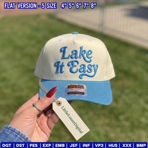 Könnte beinhalten: Cremefarbene und hellblaue Baseballkappe mit dem gestickten Text "Lake It Easy". Die Kappe hat einen gebogenen Schirm und ein Etikett mit der Aufschrift "LittleLoomDigital". Erhältlich in den Größen 10.16cm, 12.7cm, 15.24cm, 17.78cm und 20.32cm.