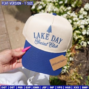 May include: A baseball cap with a cream-colored crown and a blue brim. The cap features the embroidered text "LAKE DAY Social Club" with a tree graphic. A small tag with the text "LittleLoomDigital" is attached.