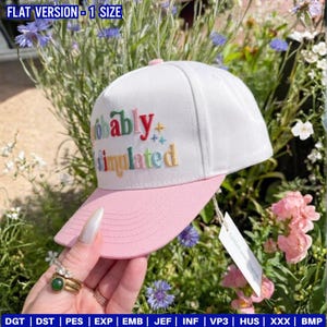 May include: A white baseball cap with a pink brim and top button. The cap features the embroidered text "Probably Simulated" in various colors. The cap is displayed outdoors with flowers in the background.