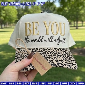 May include: A cream-colored baseball cap featuring the embroidered gold text BE YOU and the black script text the world will adjust. The brim has a black and tan leopard print pattern. A brown paper tag is attached with a piece of twine.