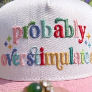 May include: White and pink baseball cap with the embroidered text "Probably overstimulated" in colorful letters. The hat has a curved brim and a pink button on top. The design includes small star and diamond accents.