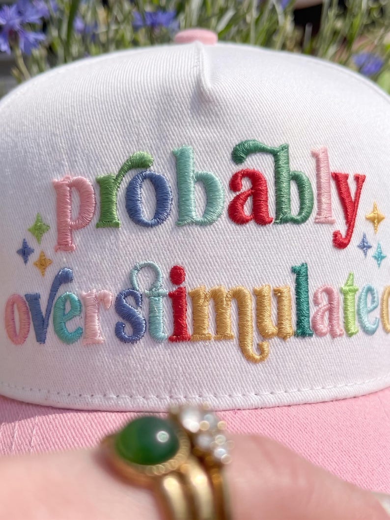 May include: White and pink baseball cap with the embroidered text "Probably overstimulated" in colorful letters. The hat has a curved brim and a pink button on top. The design includes small star and diamond accents.