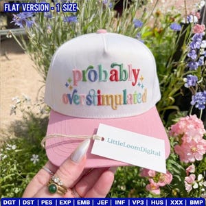 May include: A white and pink baseball cap with the embroidered phrase "probably overstimulated" in colorful letters. The cap has a curved brim and a tag that reads "LittleLoomDigital".