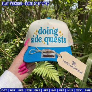 May include: A white and blue trucker hat with the phrase "doing side quests" embroidered in blue. The hat has a mesh back and a blue brim. A tag that says "LittleLoomDigital" is attached.
