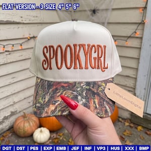 May include: A cream-colored baseball cap with the text SPOOKYGRL embroidered in orange on the front. The brim features a camouflage pattern in shades of brown, green, and black. A brown paper tag labeled LittleLoomDigital is attached to the brim.