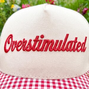 Puede incluir: Gorra de b&eacute;isbol color crema con la palabra "Overstimulated" bordada en rojo. La visera presenta un patr&oacute;n de cuadros rojos y blancos. El fondo muestra flores coloridas borrosas. Un accesorio casual.
