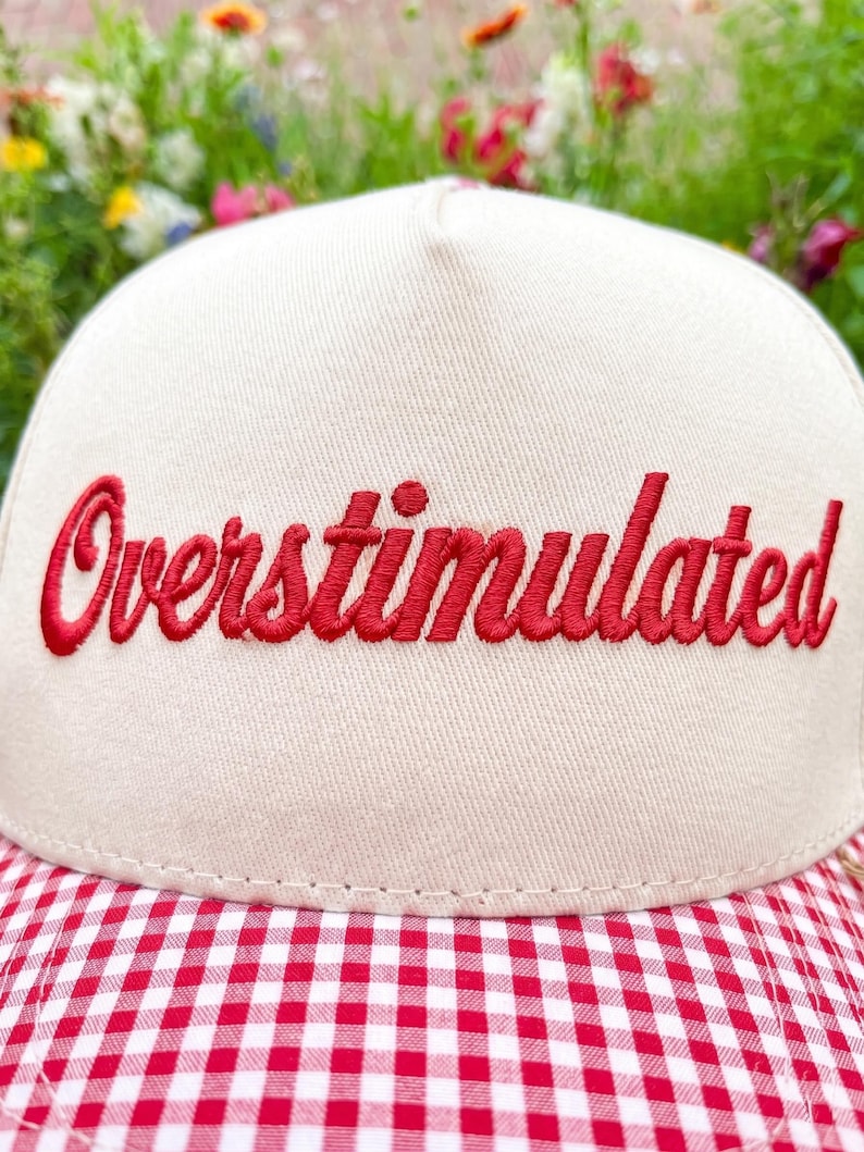 Puede incluir: Gorra de b&eacute;isbol color crema con la palabra "Overstimulated" bordada en rojo. La visera presenta un patr&oacute;n de cuadros rojos y blancos. El fondo muestra flores coloridas borrosas. Un accesorio casual.