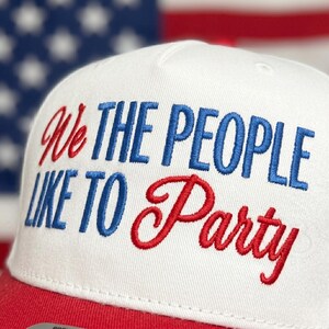May include: White and red baseball cap with the embroidered phrase "We The People Like To Party" in blue and red. The cap has a red brim and a white crown, with the American flag in the background.