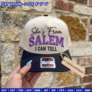 May include: A cream-coloured baseball cap with a black brim featuring the embroidered text She's From SALEM I CAN TELL in black and purple. The cap includes an Otto brand sticker on the brim and a tan tag labelled LittleLoomDigital attached.