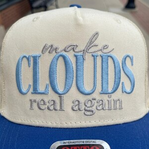 May include: A cream and blue baseball cap with the embroidered phrase "make CLOUDS real again." The word "CLOUDS" is in light blue, and the rest of the text is in gray. The cap has a blue brim.