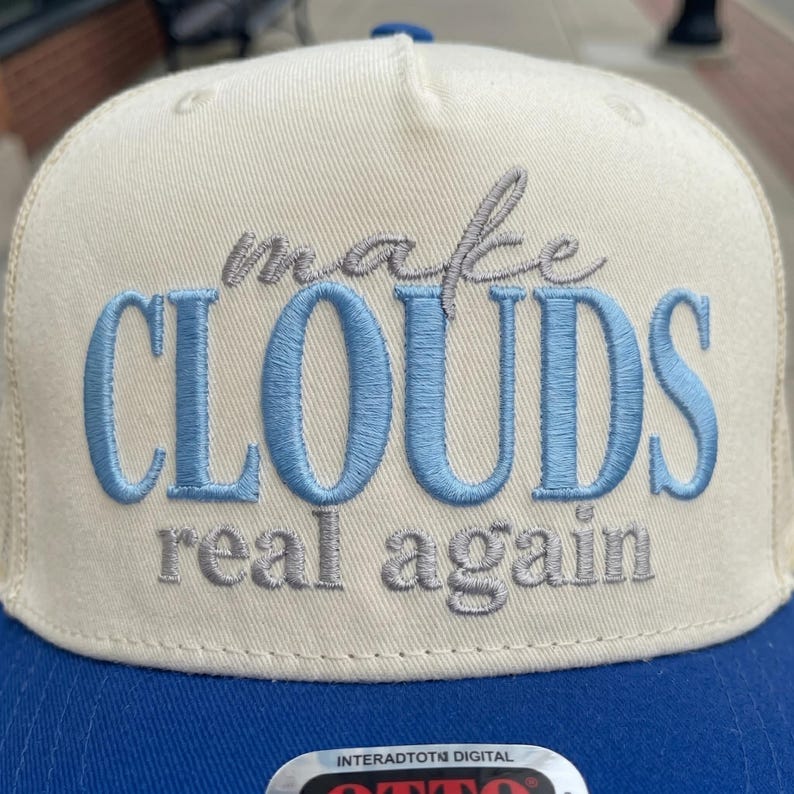 May include: A cream and blue baseball cap with the embroidered phrase "make CLOUDS real again." The word "CLOUDS" is in light blue, and the rest of the text is in gray. The cap has a blue brim.