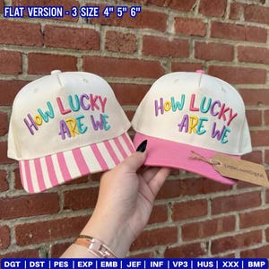 May include: Two white baseball caps with the text "How Lucky Are We" embroidered in colorful letters. One cap has a pink striped brim, the other has a solid pink brim. The caps are displayed against a brick wall.