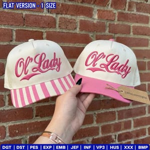 May include: Two white baseball caps with pink embroidery that reads "Ol' Lady". One cap has a pink brim and the other has a striped pink and white brim. The caps are displayed against a brick wall.