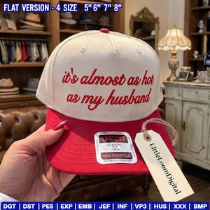 May include: A baseball cap with a cream-coloured crown and a red brim. The cap features the embroidered phrase "it's almost as hot as my husband" in red. A tag with the text "LittleLoomDigital" hangs from the cap.