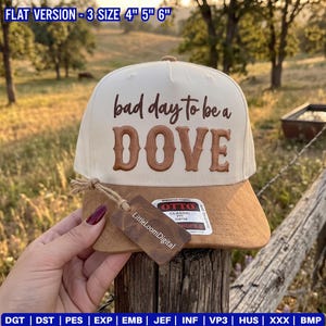May include: A cream-colored baseball cap with a brown suede-like brim. The front features the embroidered text 'bad day to be a DOVE' in brown. A brown rectangular tag labeled 'LittleLoomDigital' is attached to the brim with a jute string.