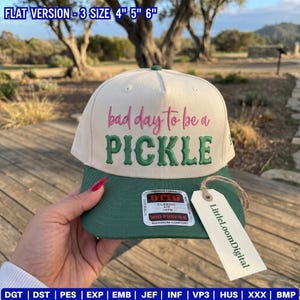 Op de afbeelding: Een crème en groene baseballcap met de tekst "bad day to be a PICKLE" geborduurd op de voorkant. De cap heeft een groene klep en een label met "LittleLoomDigital".
