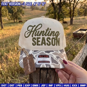 Puede incluir: Una gorra de béisbol color crema con visera de camuflaje y el texto bordado Hunting SEASON en verde oliva. La gorra presenta una pegatina de la marca OTTO en la parte delantera y una etiqueta rectangular marrón sujeta con un cordel.