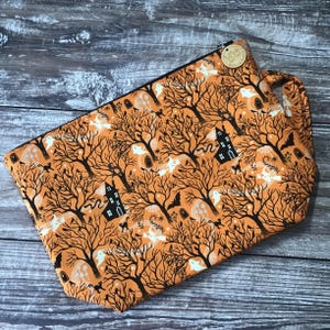 May include: Orange Halloween-themed zippered pouch with a black zip and handle. The fabric features a repeating pattern of black bare trees, ghosts, bats, and haunted houses. A small wooden tag is attached to the handle.