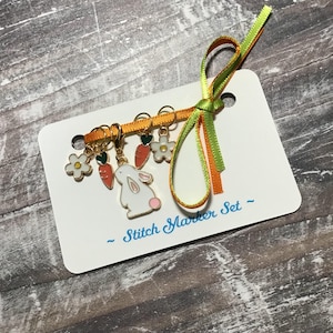 May include: A set of stitch markers on a white card, featuring a bunny, carrots, and flowers. The markers are attached to a gold-colored bar and tied with green and orange ribbons. The card reads "Stitch Marker Set".