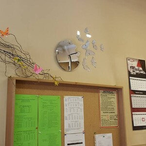 May include: A silver clock with a mirrored face and butterfly accents on a white wall. The clock is mounted on a cork board with papers and a sign with the word "INSTRUCTIONS" on it.