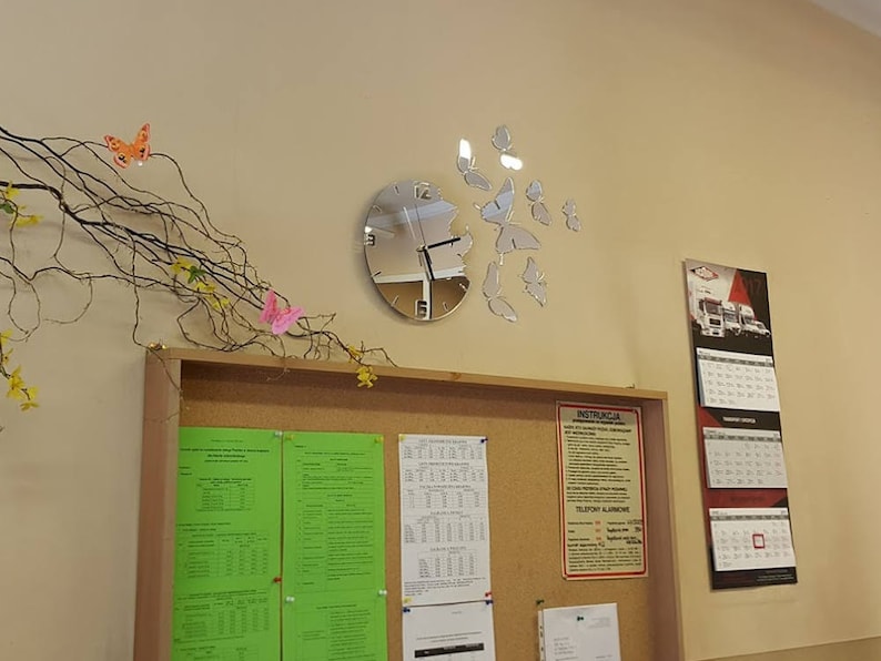 May include: A silver clock with a mirrored face and butterfly accents on a white wall. The clock is mounted on a cork board with papers and a sign with the word "INSTRUCTIONS" on it.
