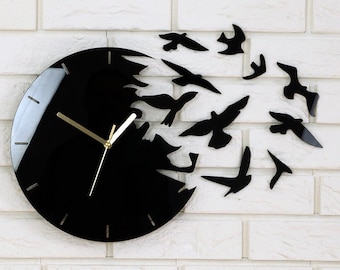 May include: A black clock with gold hands and a design of birds flying out of the clock face. The clock is mounted on a white brick wall.