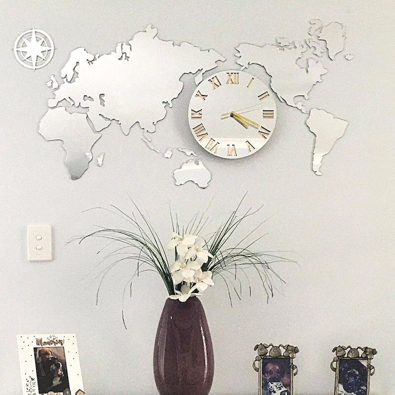 May include: A mirrored world map wall clock with Roman numeral markings on a white background. The clock is mounted on a white wall with a compass and a light switch. A vase with white flowers sits on a table in front of the clock.