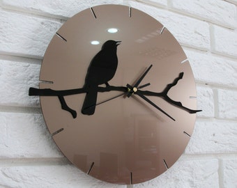 May include: A round wall clock with a metallic bronze face featuring a black silhouette of a bird perched on a branch. The clock has thin black hour and minute hands, a thin second hand, and simple black line markers for the hours.