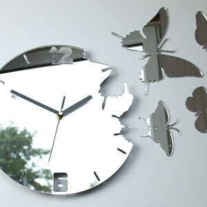 May include: A silver mirrored clock with a unique design featuring a broken edge and butterfly accents. The clock face has Roman numerals and black hands.