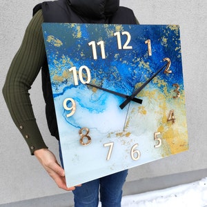 Könnte beinhalten: Quadratische Uhr mit abstraktem Design in Blau und Gold. Das Zifferblatt der Uhr hat schwarze Zeiger und goldene Zahlen.
