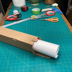 May include: A roll of white adhesive-backed vinyl with a grid pattern is partially unrolled from a cardboard box. The box is sitting on a green cutting mat with white grid lines.