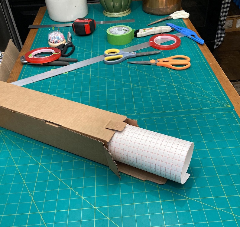 May include: A roll of white adhesive-backed vinyl with a grid pattern is partially unrolled from a cardboard box. The box is sitting on a green cutting mat with white grid lines.