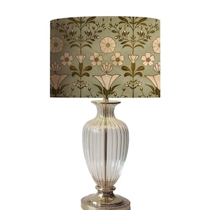 May include: A decorative table lamp with a clear, ribbed glass base and a patterned lampshade. The lampshade features a floral design in shades of green, cream, and brown. The base has a metallic finish, adding a touch of elegance to the lighting fixture.