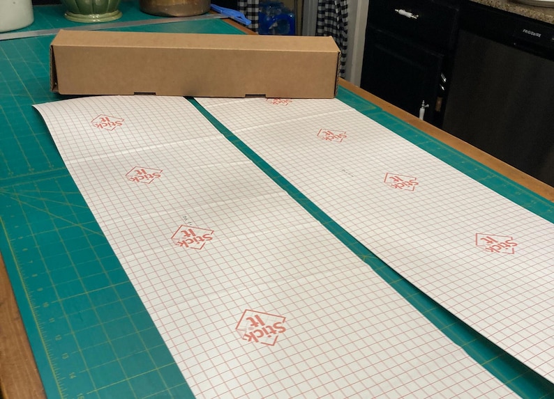 May include: Two white sheets of adhesive-backed paper with a grid pattern and the words "Stick It" printed in red on each sheet. The sheets are laid out on a green cutting mat.