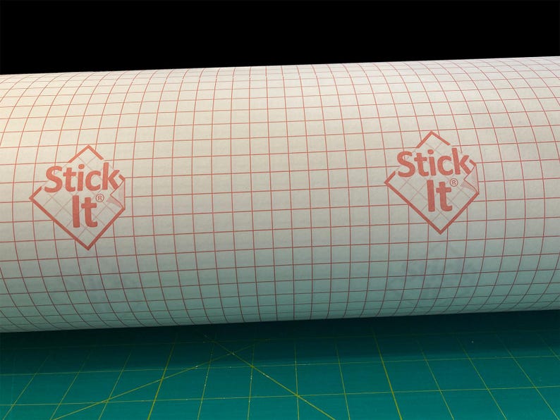May include: A roll of white adhesive-backed paper with a red grid pattern. The paper has the words "Stick It" printed on it in red.