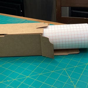 May include: A brown cardboard box with a roll of white adhesive paper with a red grid pattern inside. The box is partially open and the paper is partially unrolled.