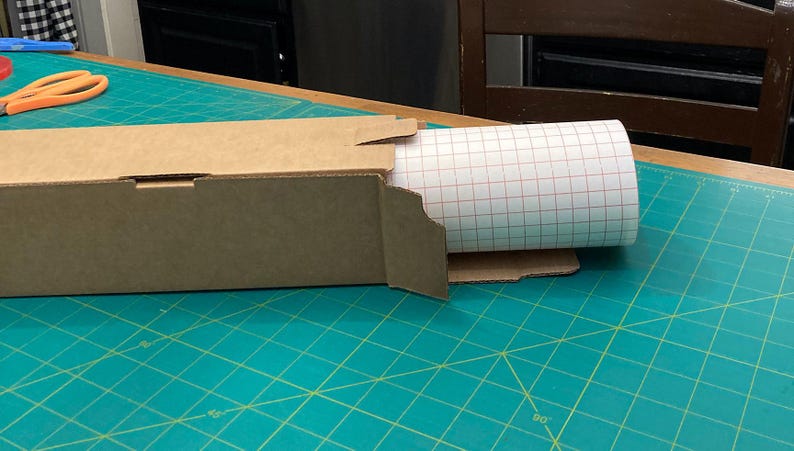 May include: A brown cardboard box with a roll of white adhesive paper with a red grid pattern inside. The box is partially open and the paper is partially unrolled.