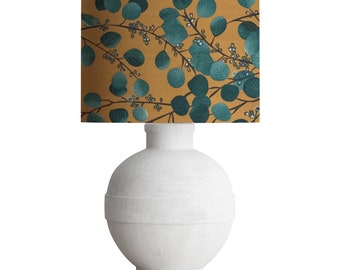 May include: A white ceramic table lamp with a round base and a cylindrical lampshade. The lampshade has a mustard yellow background with a pattern of green eucalyptus leaves.