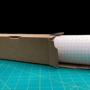 May include: A roll of white adhesive-backed paper with a red grid pattern is partially unrolled from a brown cardboard box. The box is sitting on a green cutting mat.