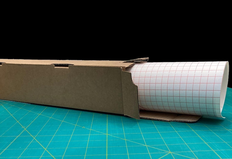 May include: A roll of white adhesive-backed paper with a red grid pattern is partially unrolled from a brown cardboard box. The box is sitting on a green cutting mat.