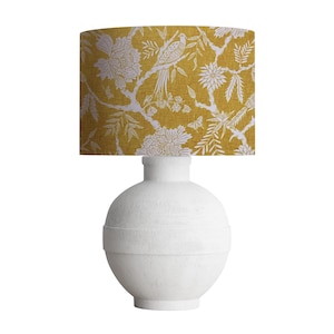 May include: A decorative table lamp featuring a white ceramic base and a mustard yellow lampshade. The lampshade is adorned with a white floral and bird pattern. The lamp has a round base and a cylindrical neck.