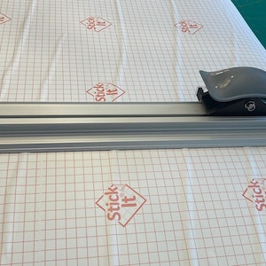 May include: A silver cutting mat with a grid pattern and the words "Stick It" printed on it. A gray plastic cutter is on the mat.